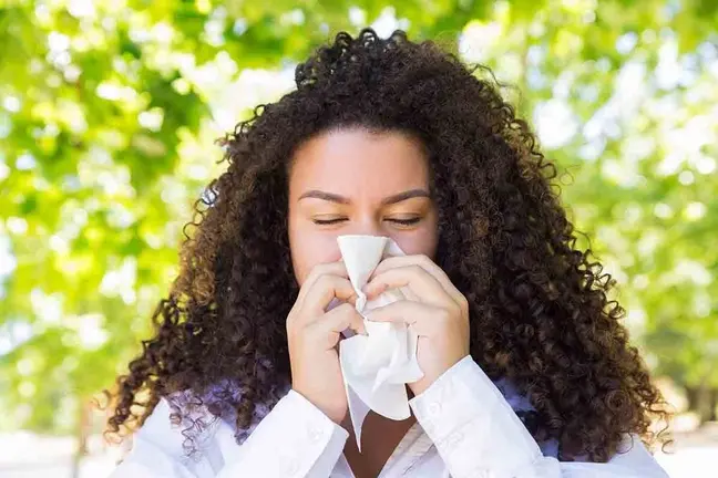 Beautiful woman blowing nose in park. Pretty young lady wearing blouse, holding handkerchief and keeping her eyes closed with green trees in background. Cold and nature concept. Front view.