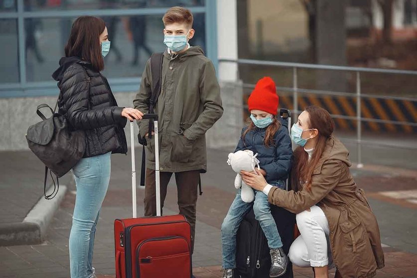 European mothers in respirators with kids are standing near a building. The travellers are protected from virus by masks.