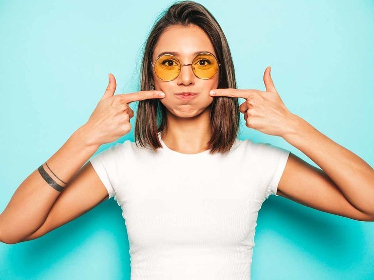 Young beautiful woman looking at camera.Trendy girl in casual summer white T-shirt and yellow skirt in round sunglasses. Positive female shows facial emotions.Funny model blowing her cheeks.Puff out