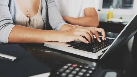 Hands of young woman working on laptop at home with husband sitting next to her