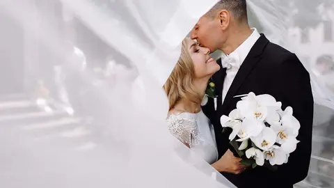 Groom in black tuxedo hugs tender stunning bride while they stand on the street of old European town