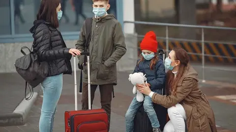 European mothers in respirators with kids are standing near a building. The travellers are protected from virus by masks.