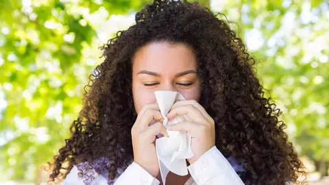 Beautiful woman blowing nose in park. Pretty young lady wearing blouse, holding handkerchief and keeping her eyes closed with green trees in background. Cold and nature concept. Front view.